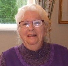 Woman with white hair and glasses smiles, wearing a purple sweater, indoors.