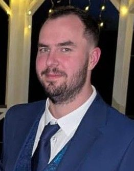 Man in a navy blue suit, white shirt, and blue tie, with a short beard, smiles, outdoors with lights.