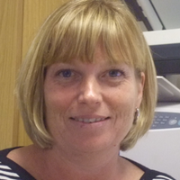 Blond woman with bob haircut, smiling. Wearing a black and white striped shirt. Sitting near a printer.