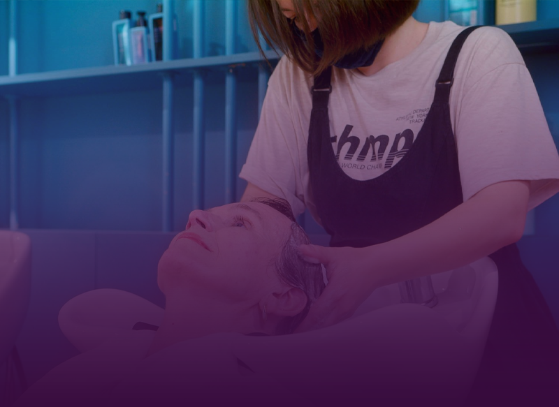 A person having their hair washed at a salon. A stylist in a black apron leans over them.