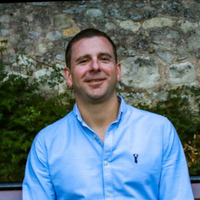 Man in a light blue button-down shirt smiles, standing in front of a stone wall with greenery.