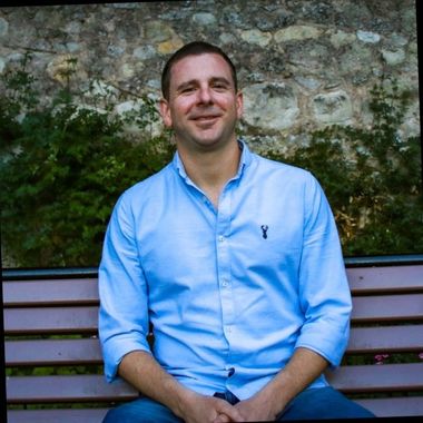 Man in blue shirt, smiling, sitting on a bench; stone wall and greenery in the background.