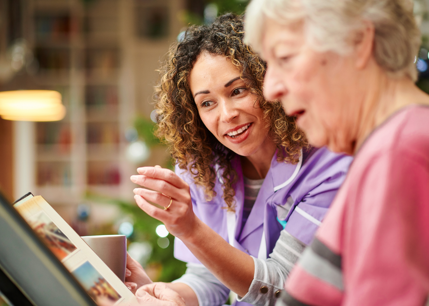 Caregiver and senior woman looking at a photo album, pointing, smiling indoors.