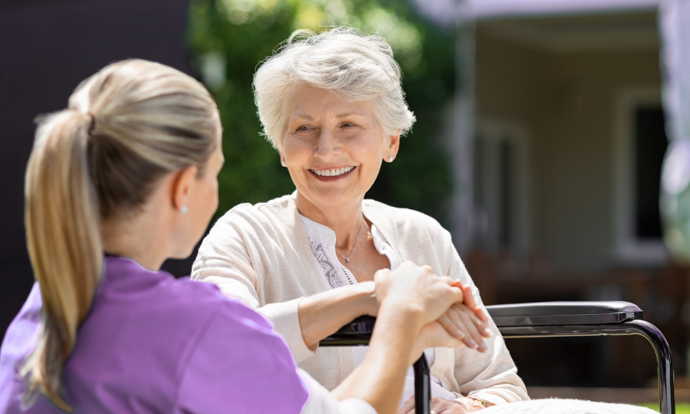 Woman in wheelchair smiles while holding hands with a caregiver, outdoors in daylight.