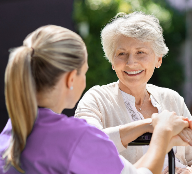 Woman in wheelchair smiles at a caregiver holding her hands outdoors.