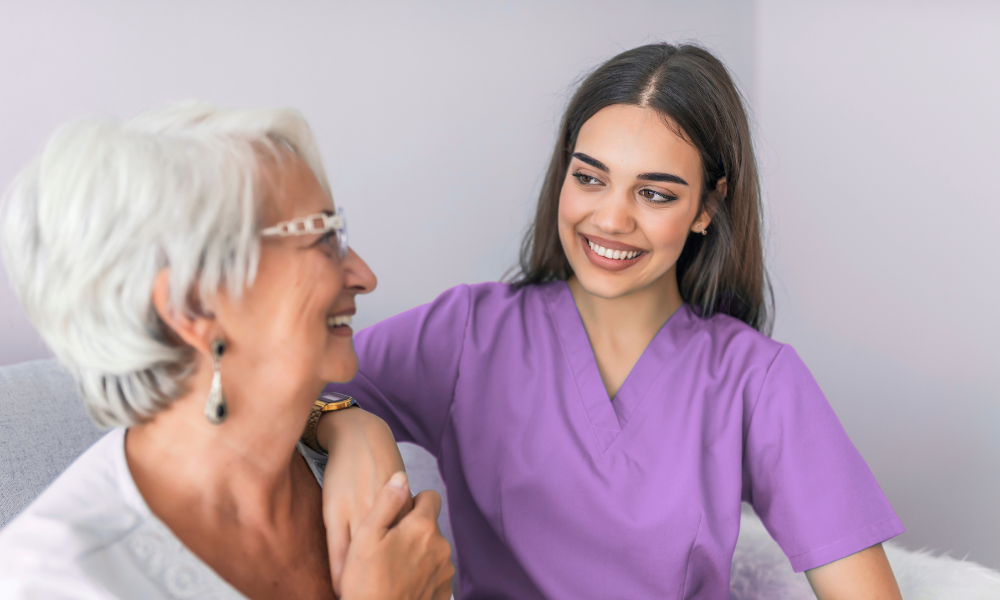 Woman in purple scrubs smiles at an older woman; both look happy. Indoors, they touch arms.