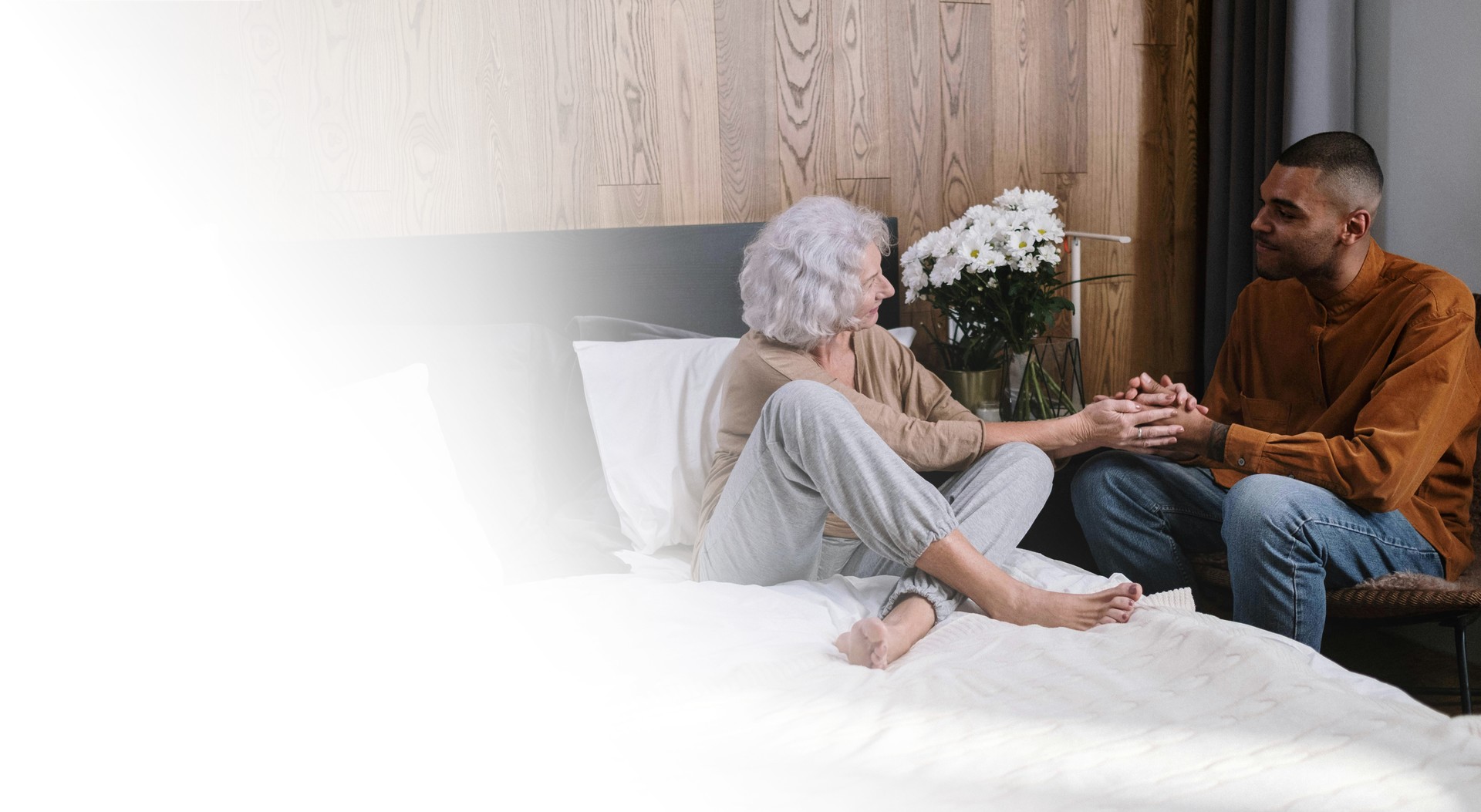 Woman and man holding hands on a bed, with flowers; soft lighting, wood paneling.