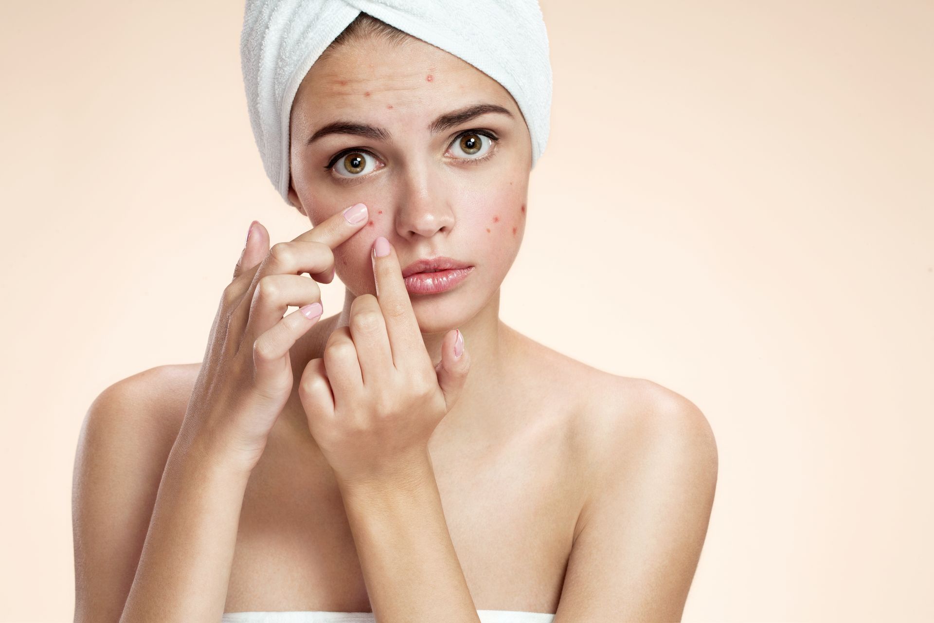 Woman in a white towel applying cream under her eye against a light background