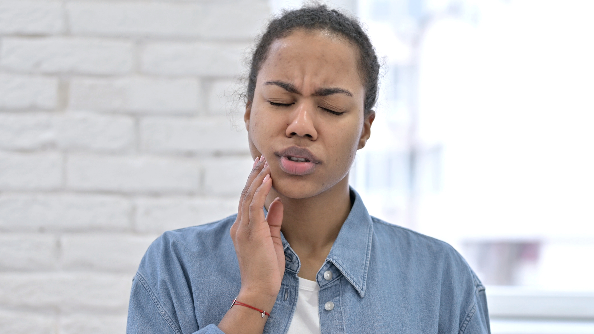 Person with eyes closed, holding cheek in pain against a light brick wall.