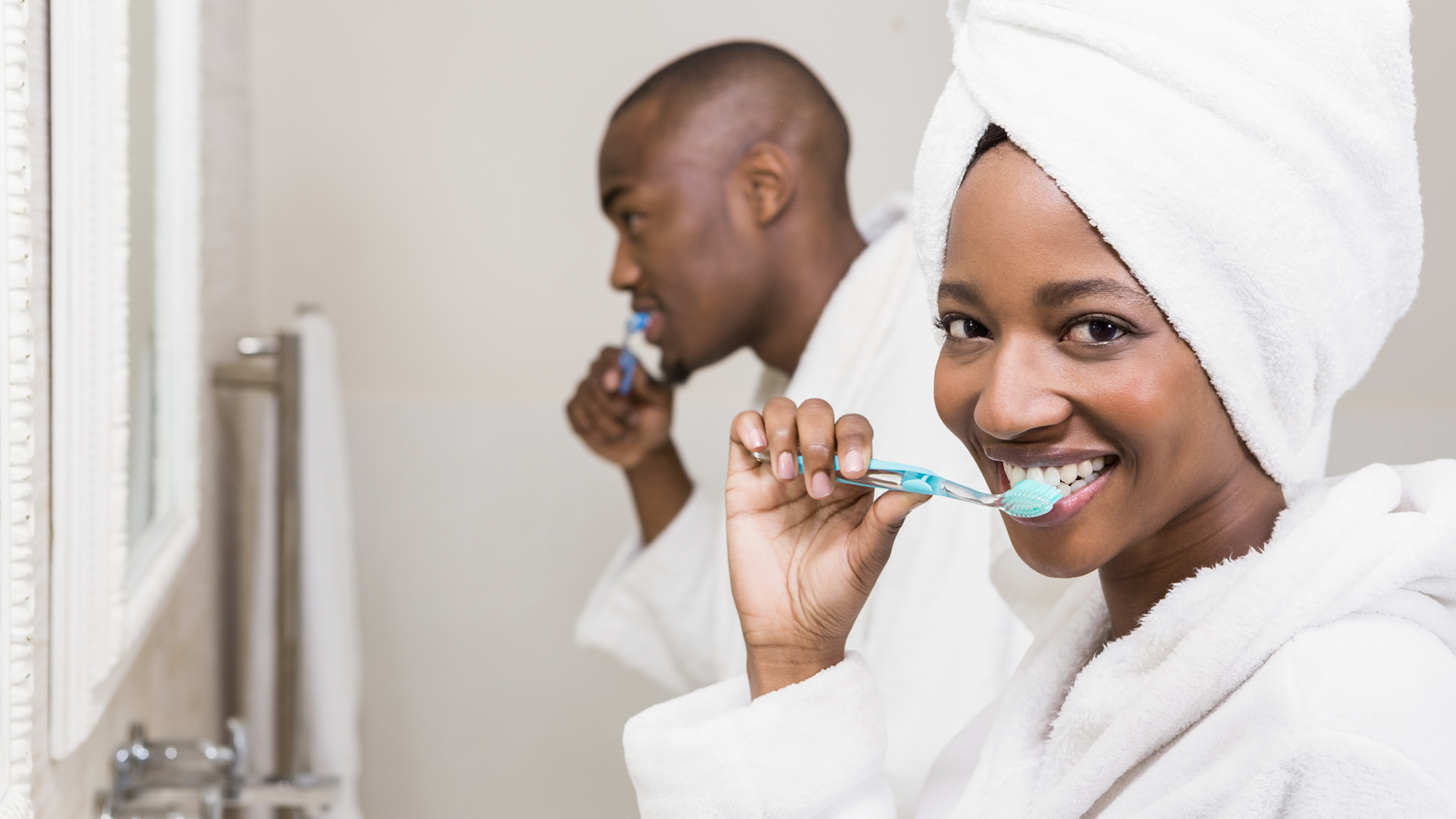 A couple brushing their teeth together in the bathroom wearing white robes.
