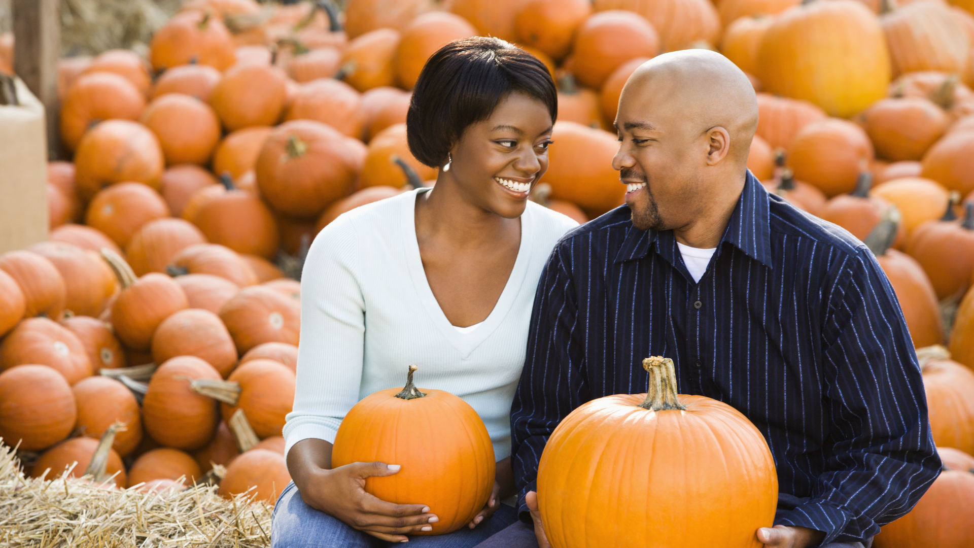 Two people smiling while holding pumpkins in a pumpkin patch with many pumpkins behind them.