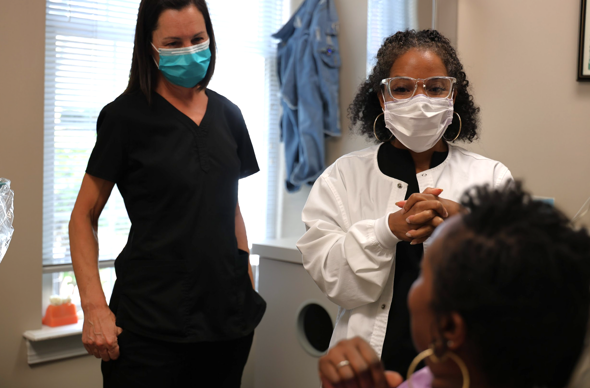 Two masked healthcare workers talking with a seated patient in a clinic room