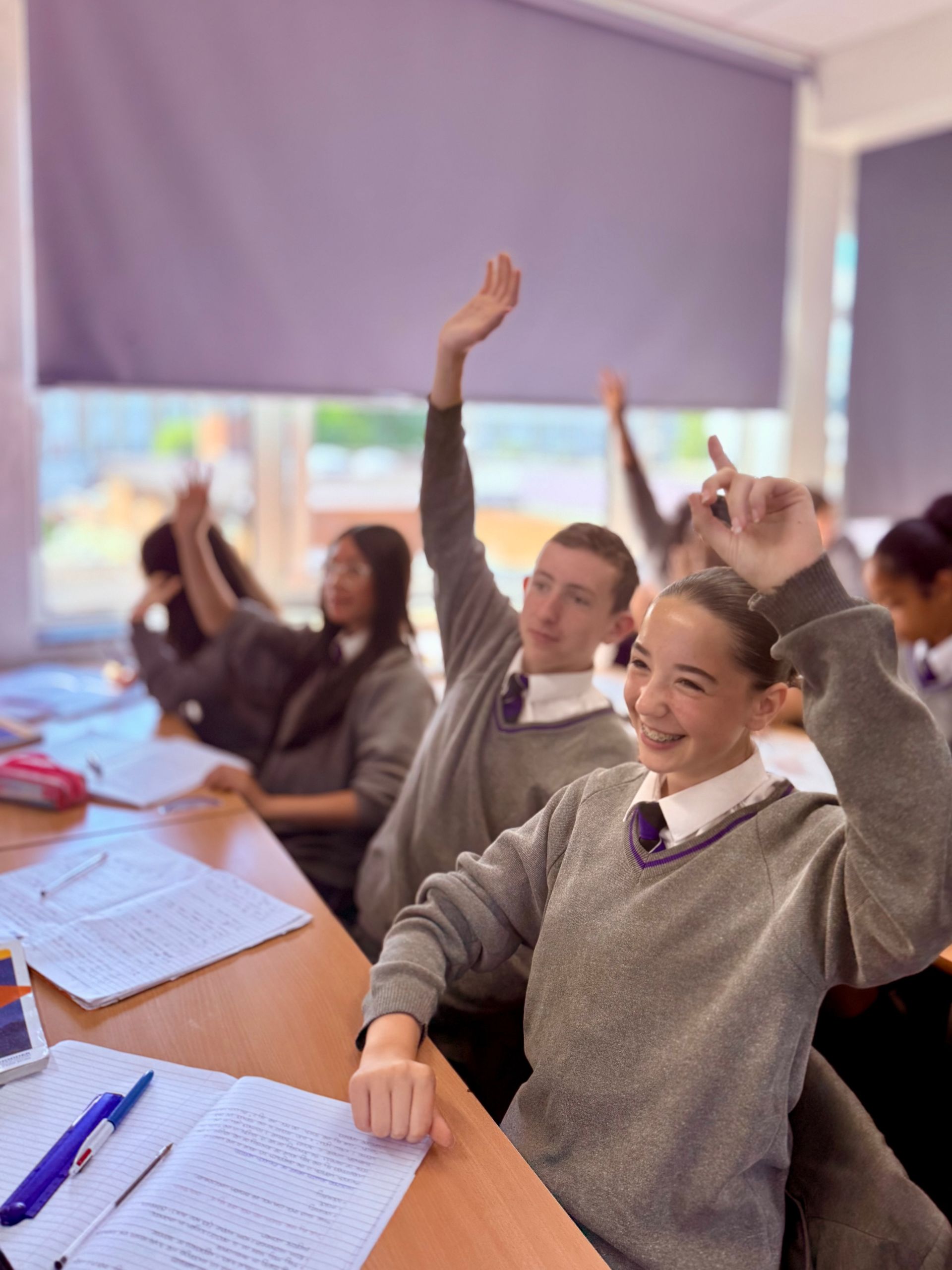 Students in gray school uniforms sitting at desks with their hands raised in a classroom.