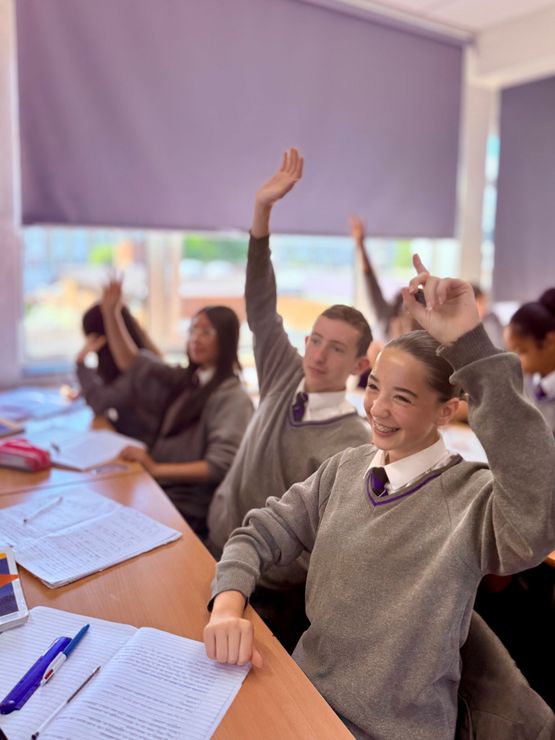 Students in gray school uniforms sitting at desks with their hands raised in a classroom.