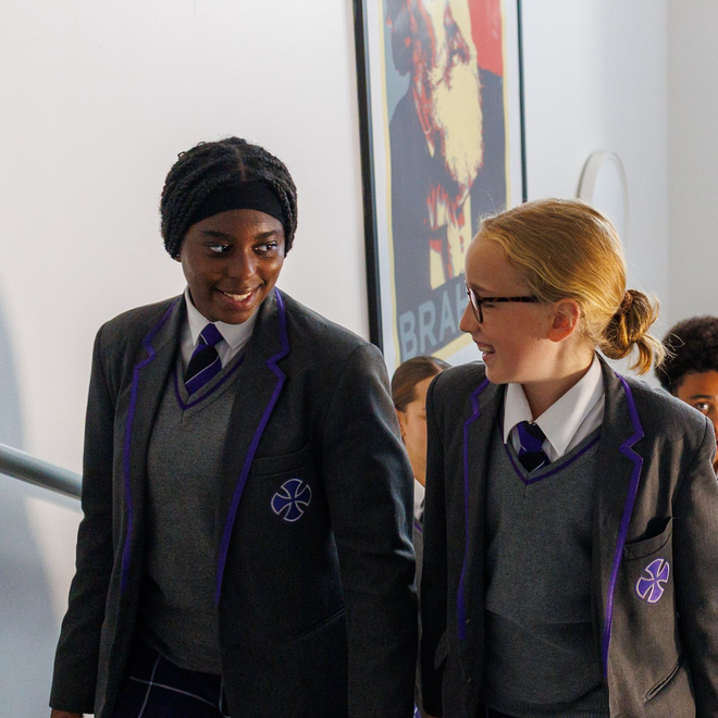 Two students in school uniforms holding certificates in a hallway, smiling at the camera.