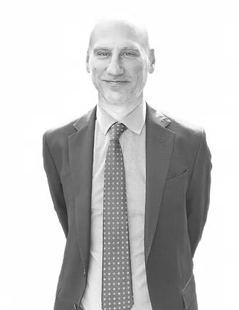 Man in a suit and tie standing against a white background, smiling slightly.