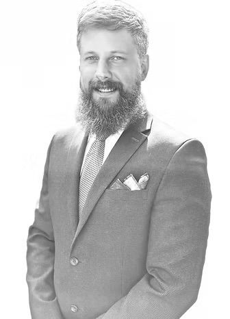 Smiling bearded man in a suit and tie with a pocket square, standing against a bright background