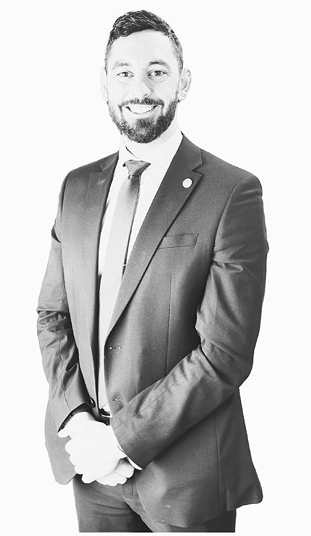 Man in a dark suit and tie standing against a plain background, wearing a lanyard and smiling.