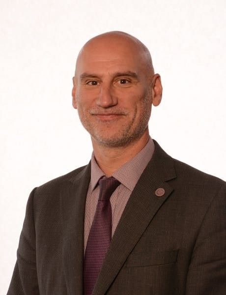Headshot of a bald man in a dark suit and purple tie, smiling against a light background