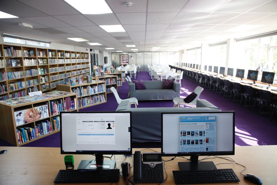 Library computer lab with dual monitors, bookshelves, and purple seating in a bright room