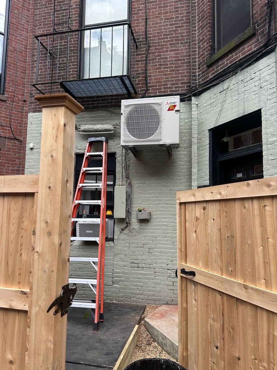 An outdoor HVAC unit mounted on a metal frame next to a wall, with a small yellow bucket on the ground nearby.