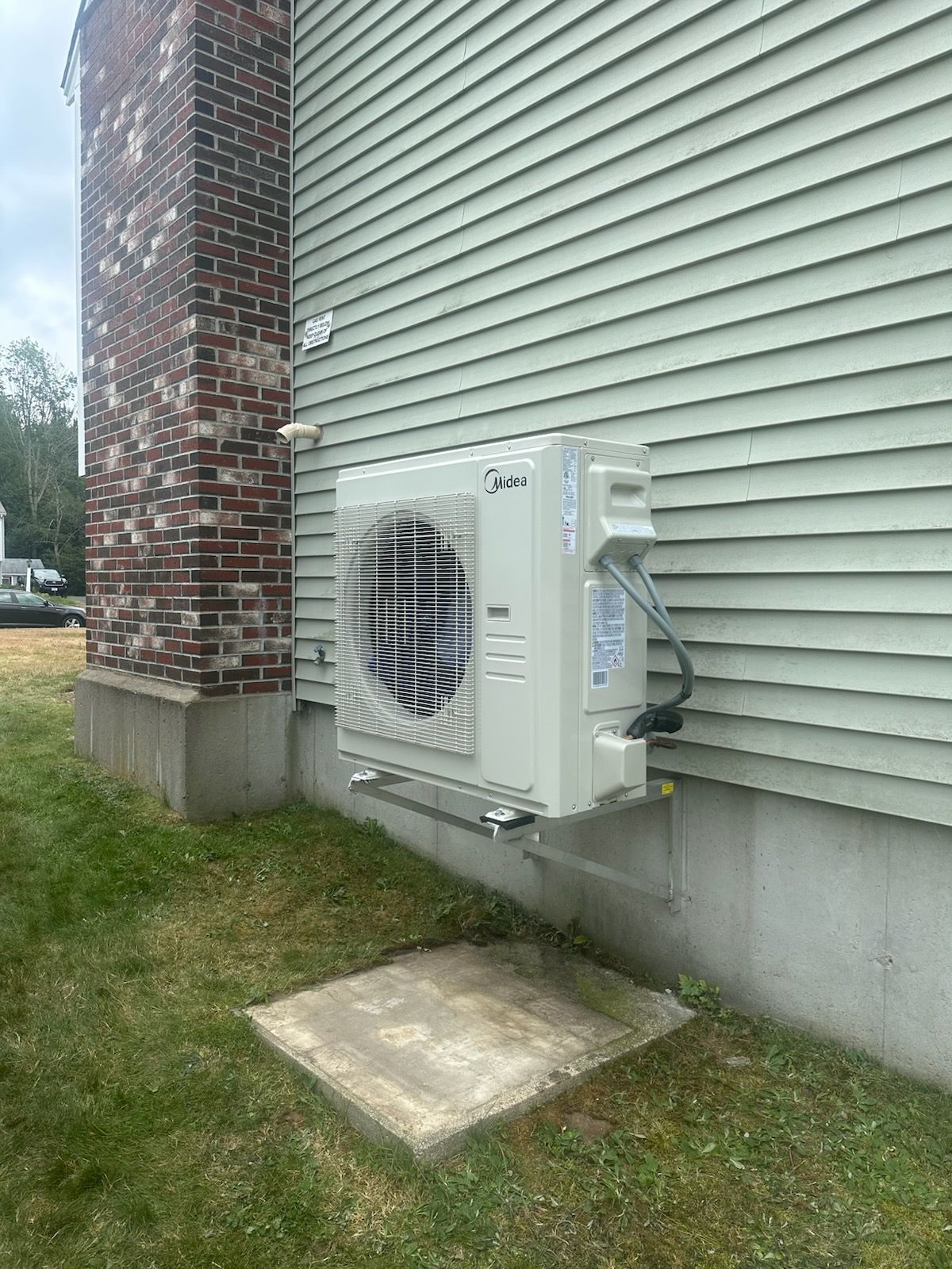 Outdoor air conditioner unit mounted on beige house siding beside a brick chimney and concrete pad.