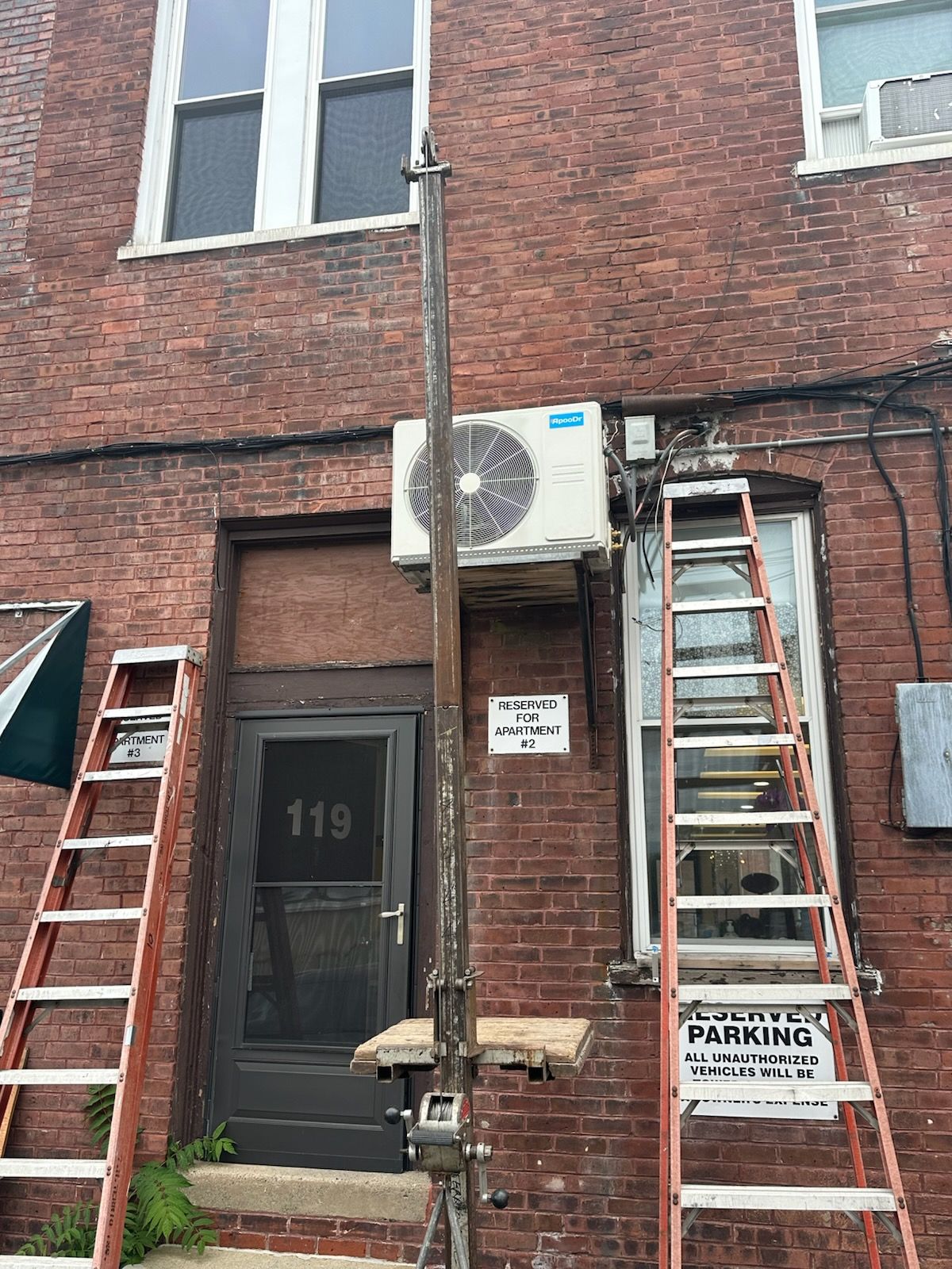 Brick building facade with two ladders, door 119, and an air conditioning unit above the entrance