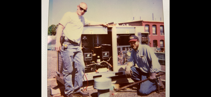 Two workers inspect machinery beside a truck outside a brick building