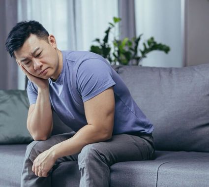 Man sitting on a couch, holding his cheek with a pained expression in a gray living room.