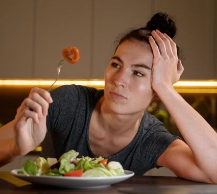 Woman at a table looking at a forkful of food, resting her head on her hand beside a salad plate.
