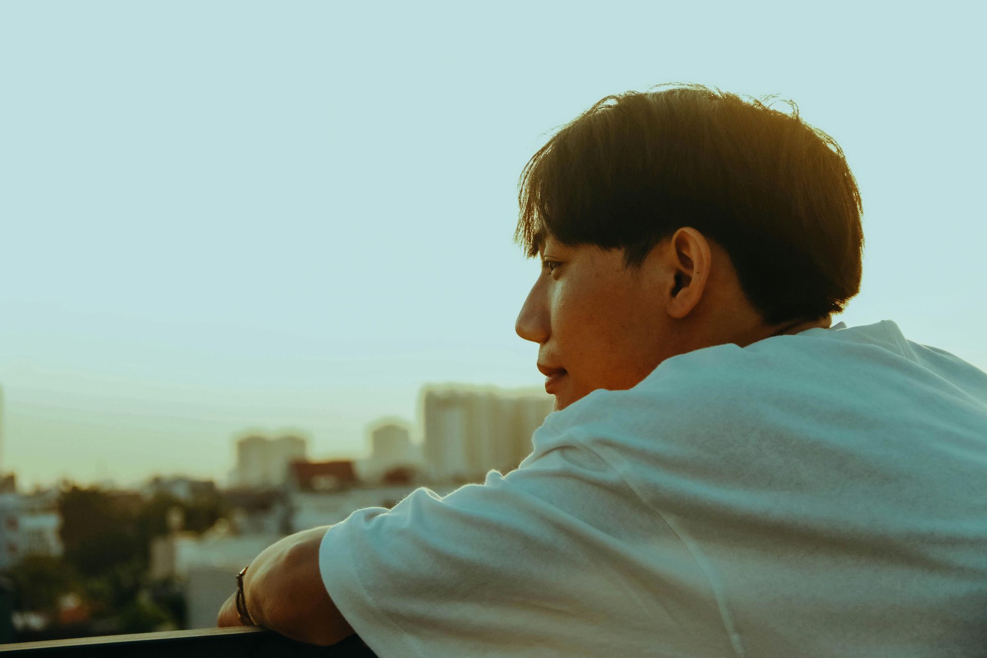 Person in a white shirt leaning on a railing, looking over a city at sunset.