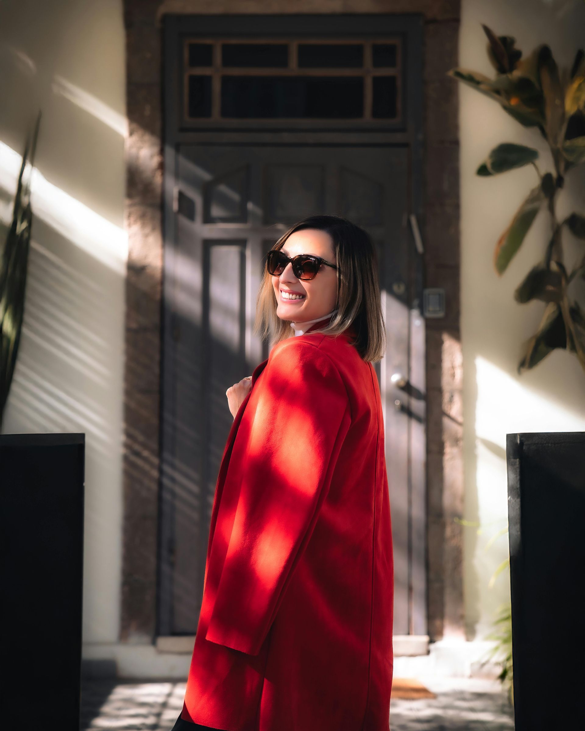 Person in a bright red coat and sunglasses standing in sunlight at a doorway with shadows across the wall.