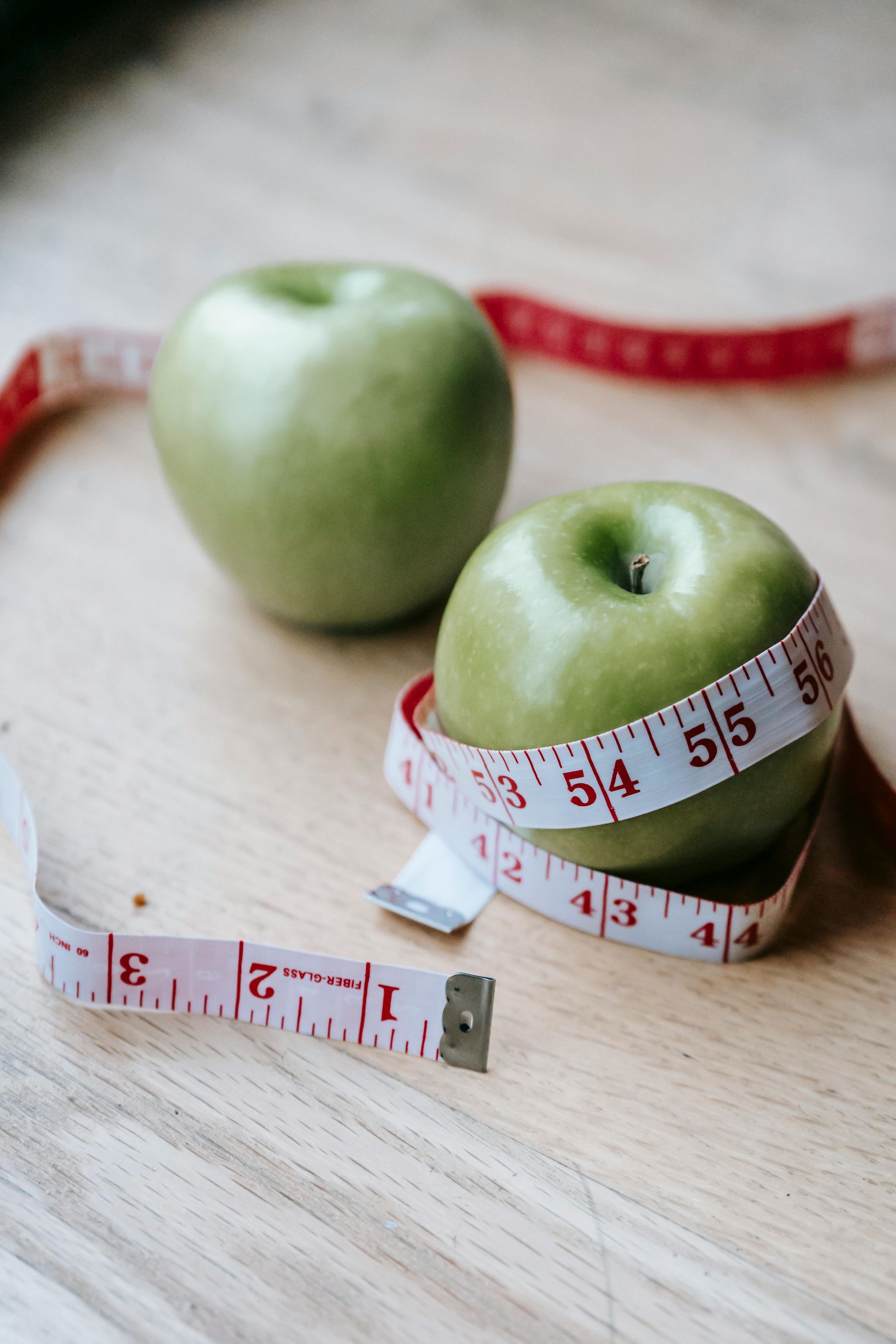 Two green apples with a measuring tape wrapped around one, on a wooden surface