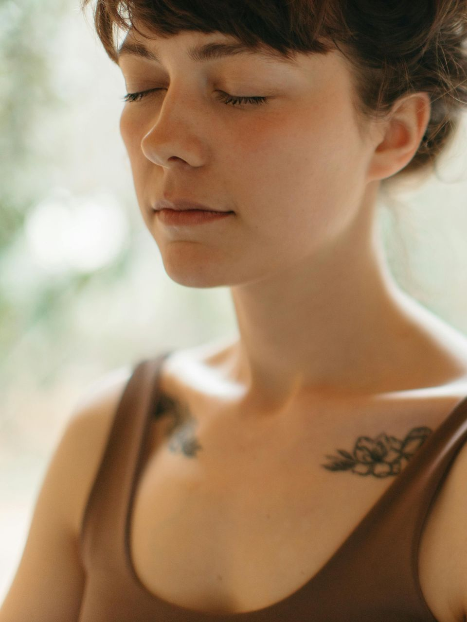 Close-up of a person with eyes closed, wearing a brown top, in soft natural light.