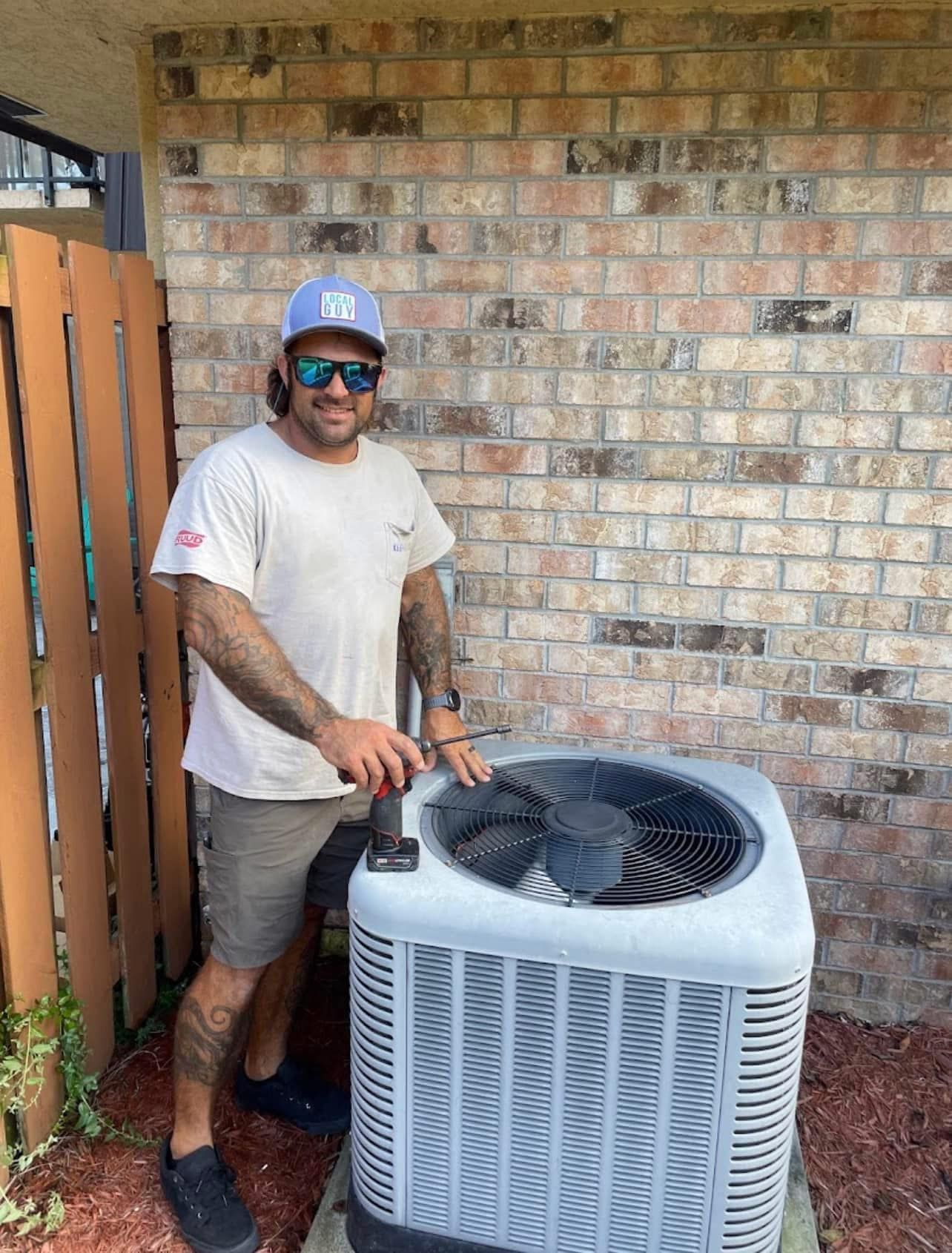 A Local Guy technician is standing next to an air conditioner in front of a brick wall.
