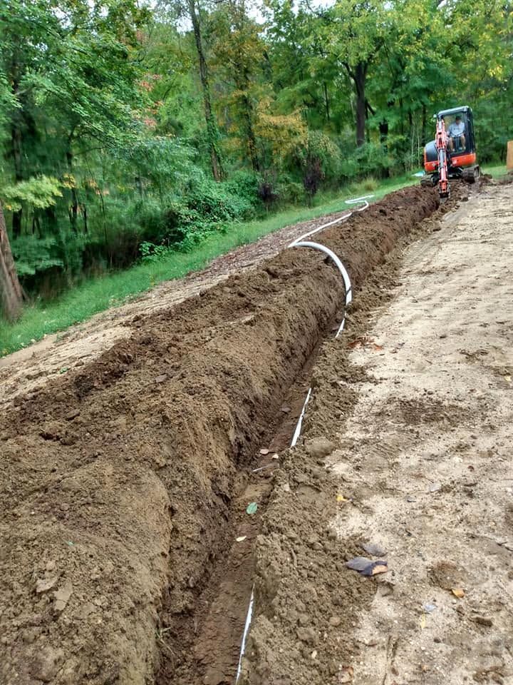 Excavator digging a long trench beside a dirt road in a wooded area
