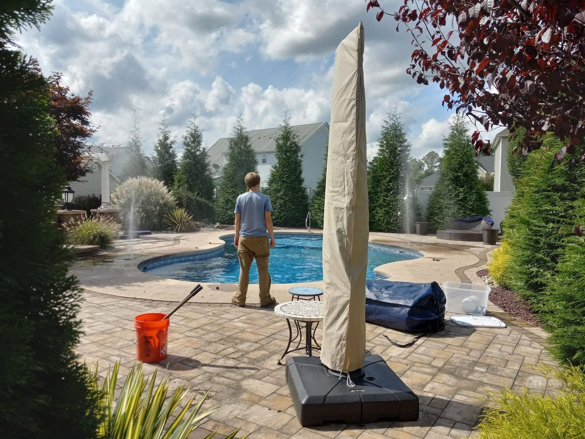 Person power-washing a pool deck beside a blue pool, with patio furniture and trees in the yard.