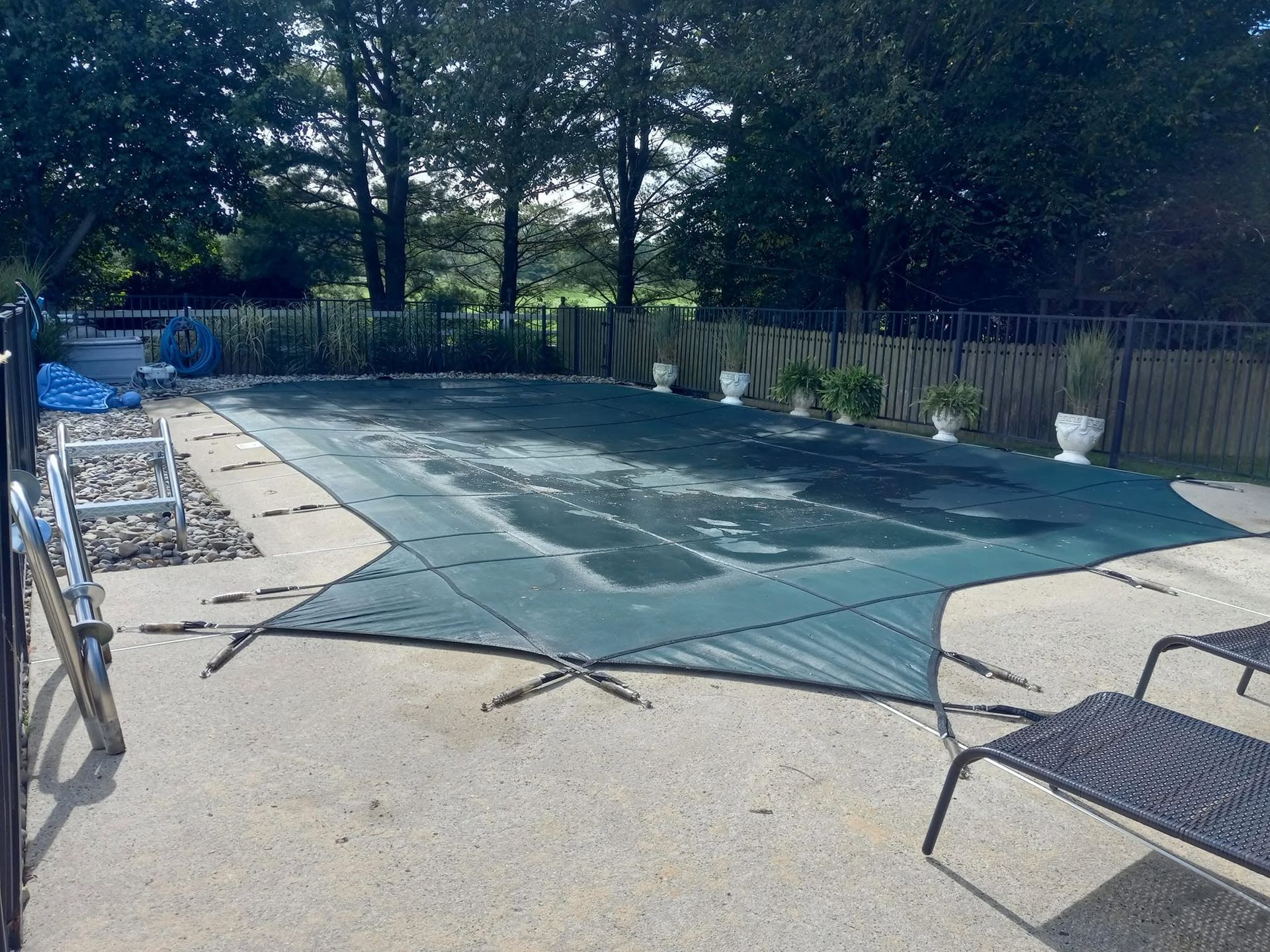 Covered outdoor swimming pool with a teal safety cover, bordered by a patio and trees.