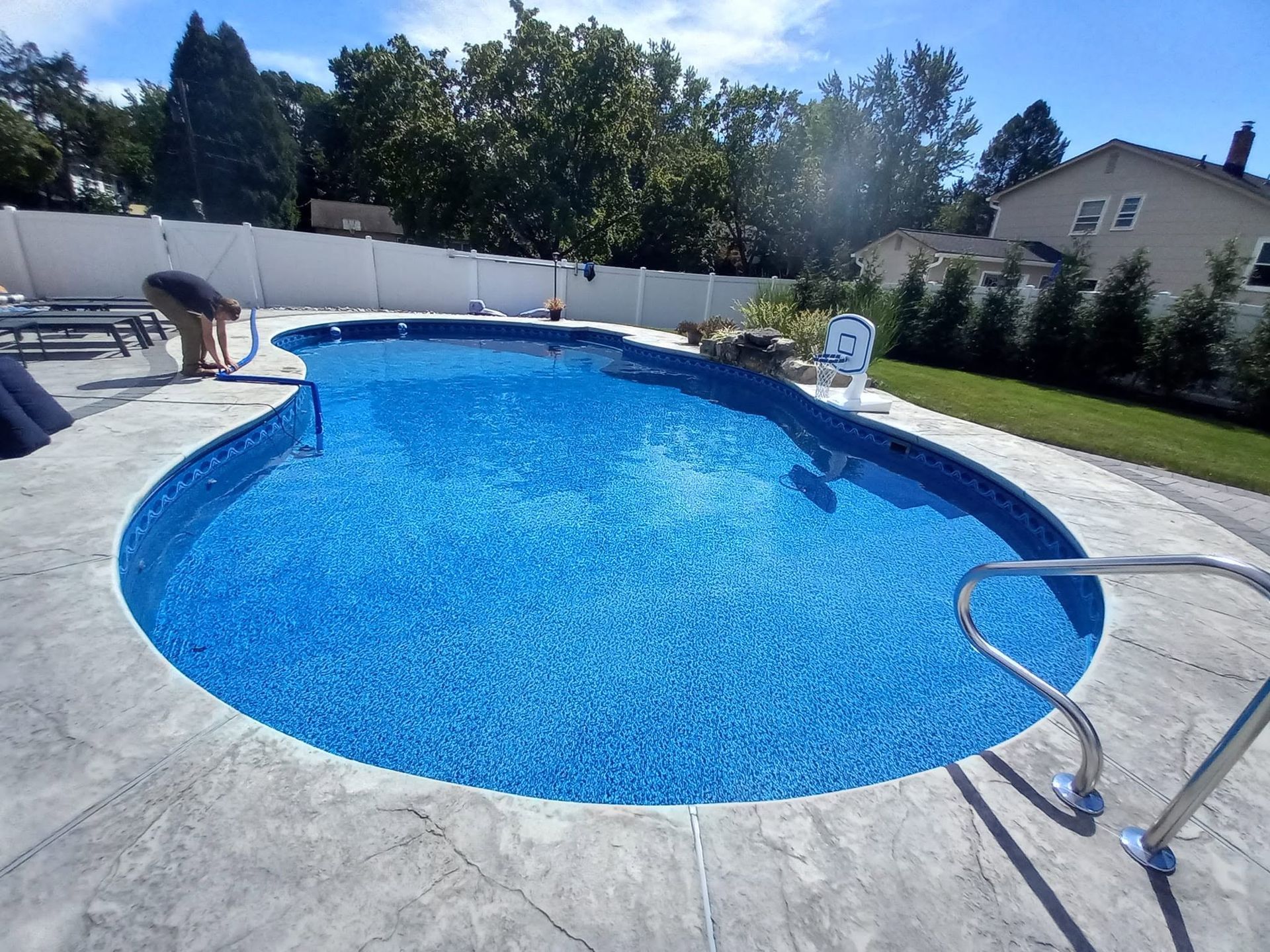 Backyard swimming pool with clear blue water, patio, fence, and trees in bright sunlight