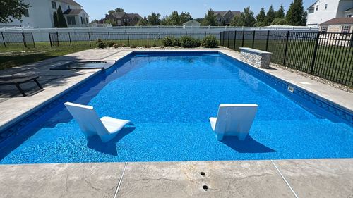 Backyard swimming pool with blue water, concrete deck, fence, and trees in bright sunlight