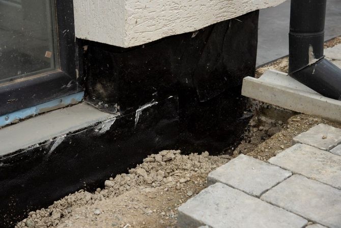 Close-up of a building corner with black waterproofing, gravel, and gray paving stones beside a window.