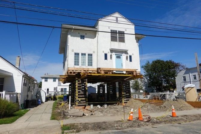 House lifted on stilts during foundation repair beside a street with orange cones and exposed ground