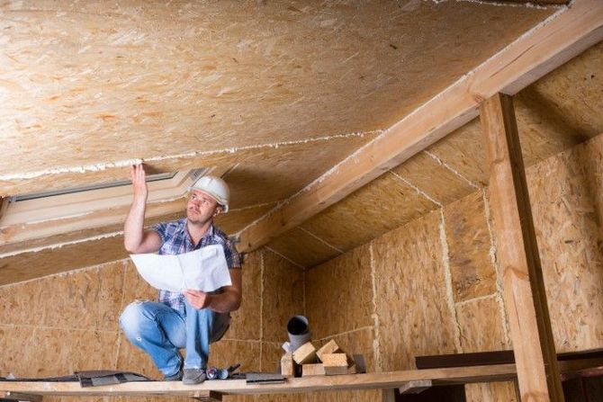 Worker installing insulation in an attic, kneeling under a sloped wooden roof with tools nearby.