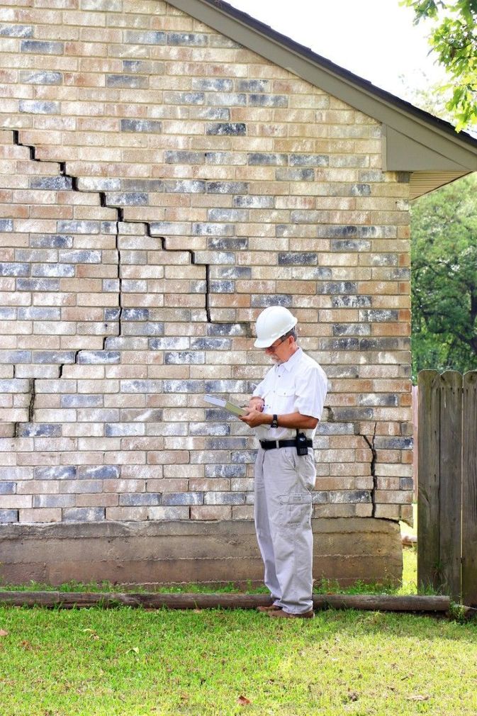 Inspector in white hard hat examines cracked brick wall beside a house.