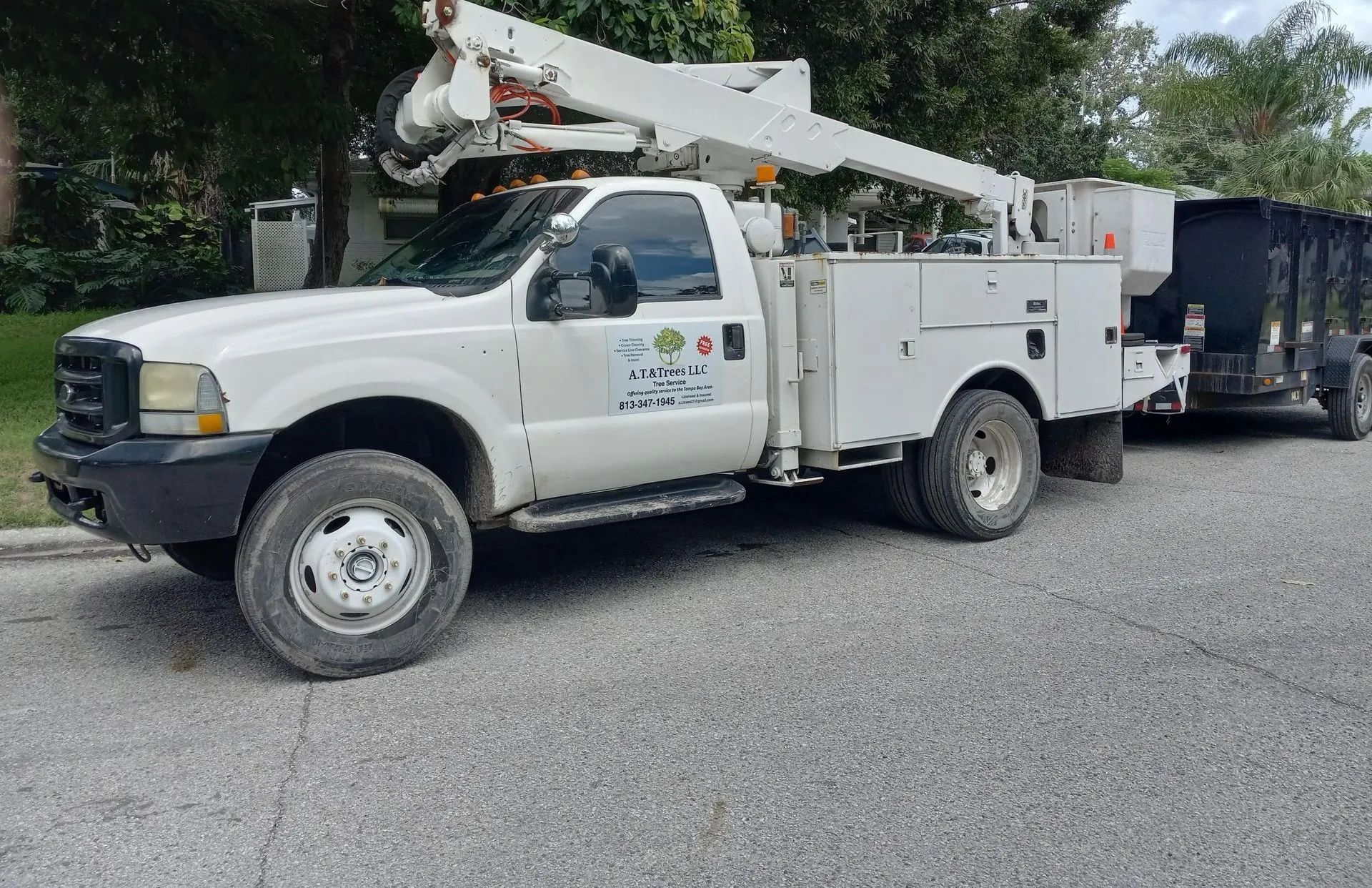 White utility truck with boom lift parked on gravel beside a black trailer and trees