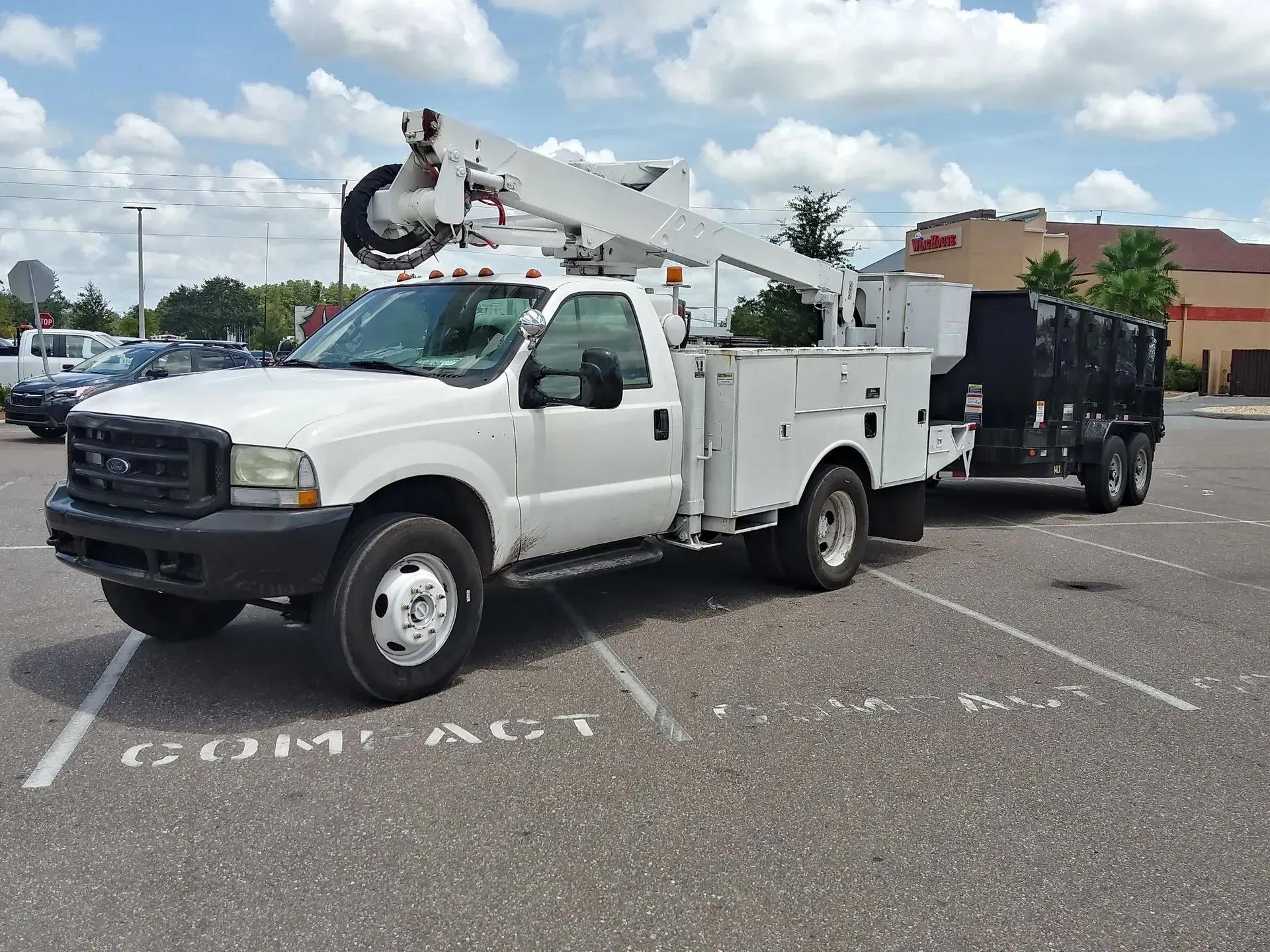 White utility truck with a boom lift parked in a lot, towing a black trailer under a cloudy sky