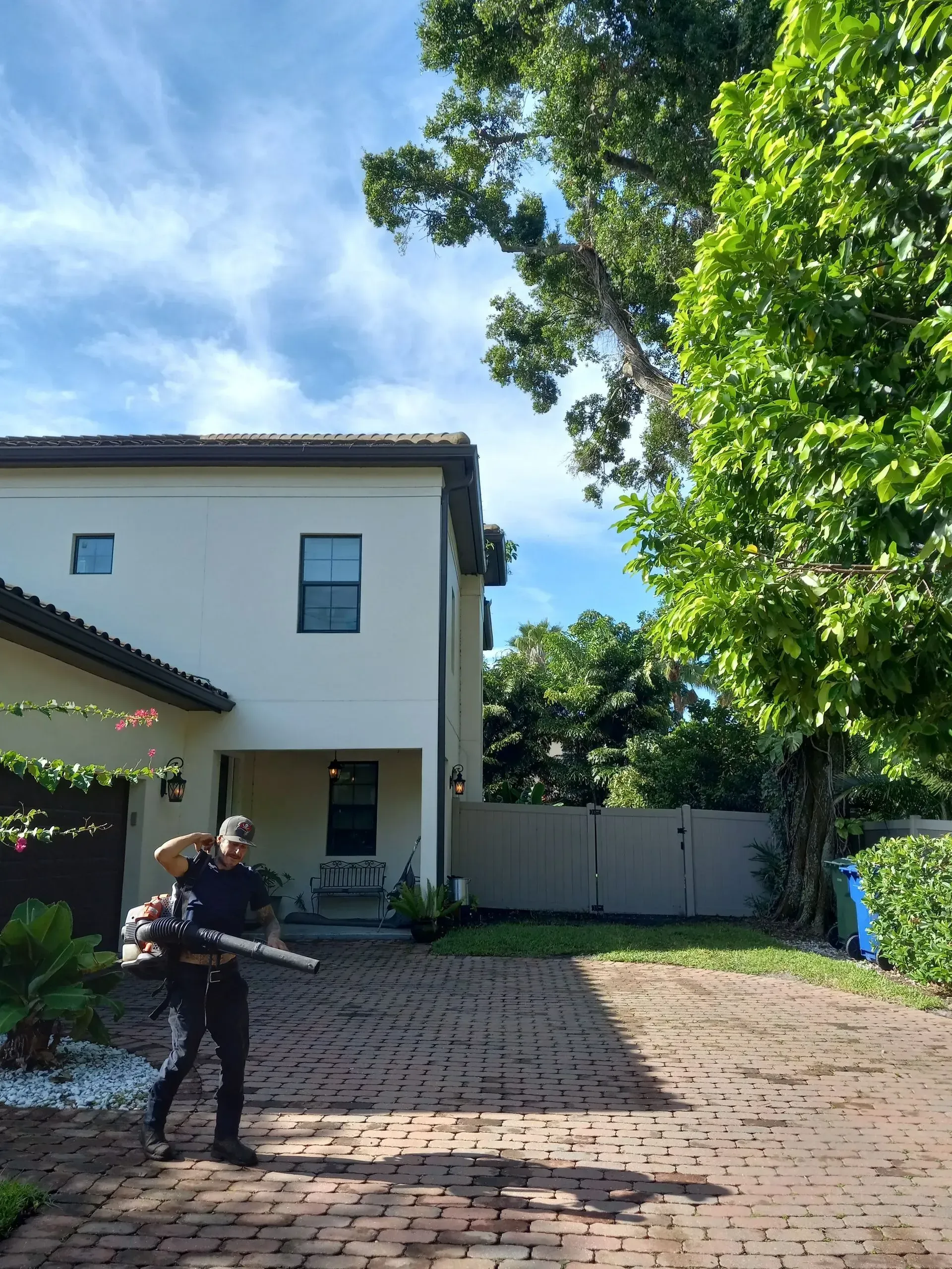 Person standing in a brick driveway beside a two-story house and trees on a sunny day
