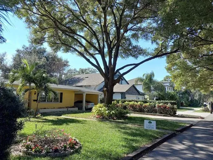 Tree-shaded suburban house with yellow exterior, green lawn, and flower beds on a sunny day