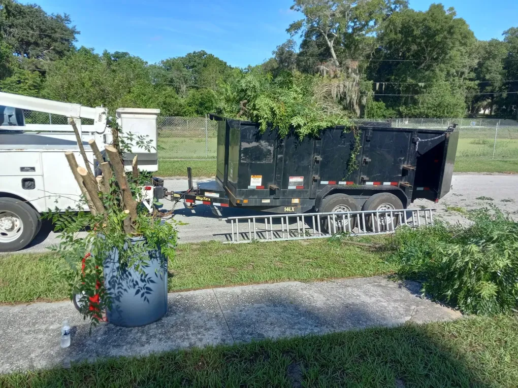 White utility trailer on a gravel lot beside a flower pot, with trees and grass in the background.