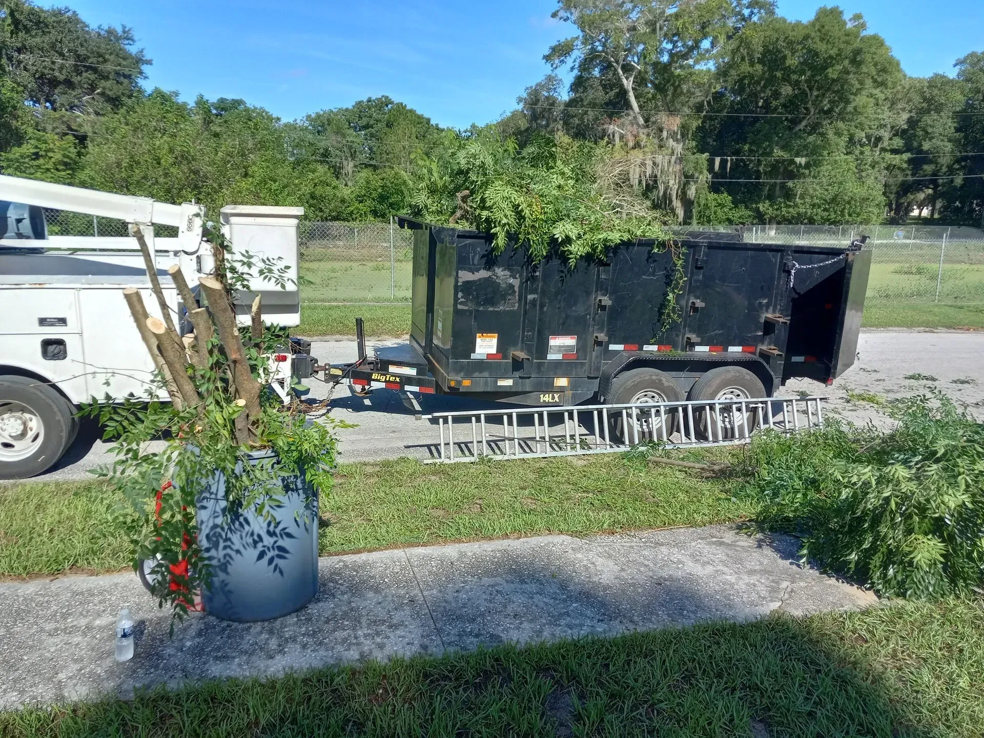 Black trailer hitched to a white truck in a grassy yard, with stacked chairs and a blue planter in front.